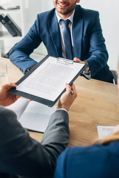 Cropped view of smiling employee holding clipboard with resume near recruiter in office 
