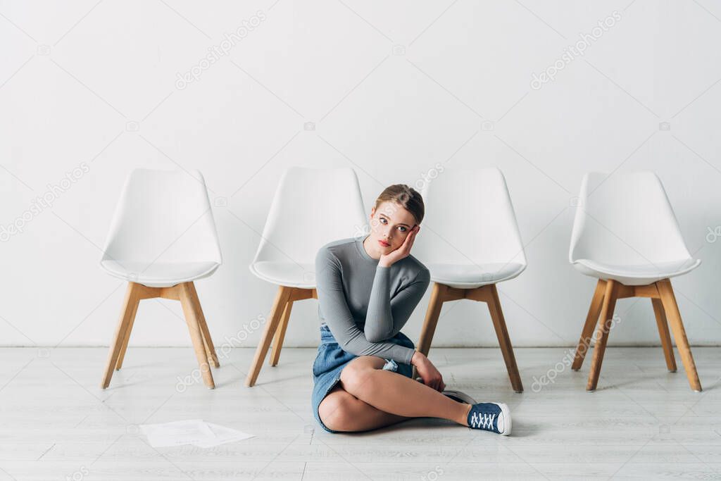 Pensive employee with hand near cheek sitting on floor near resume in office 