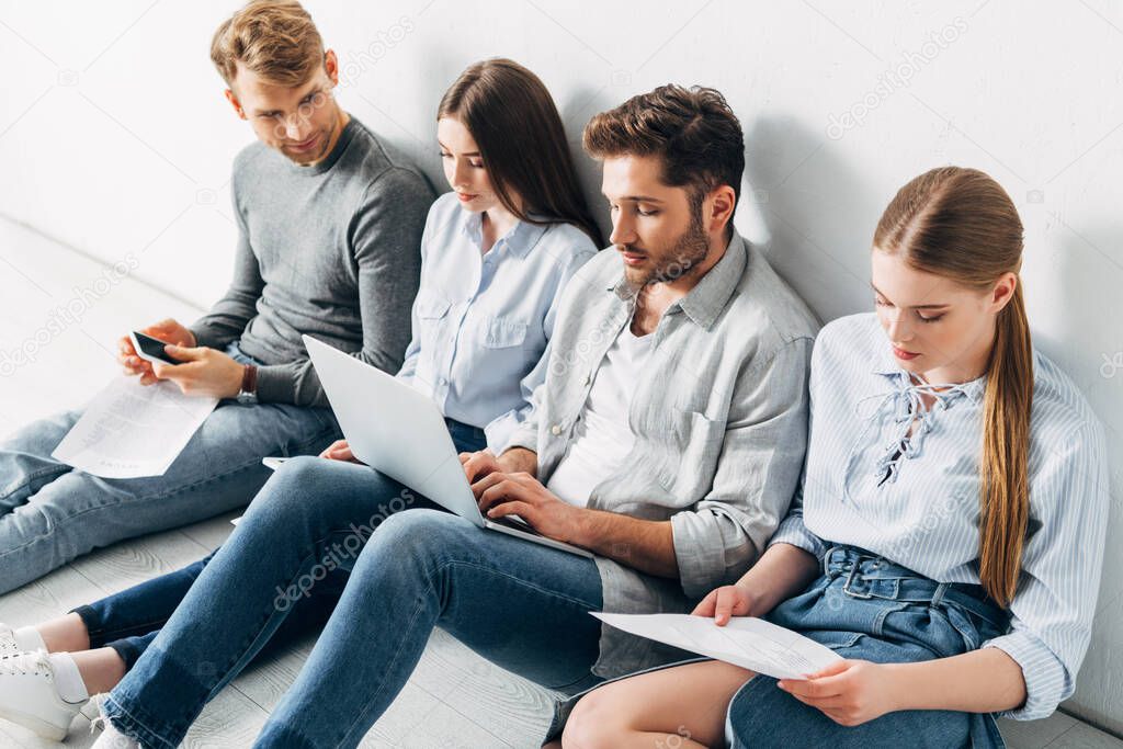 Group of young people with digital devices sitting on floor while waiting for job interview