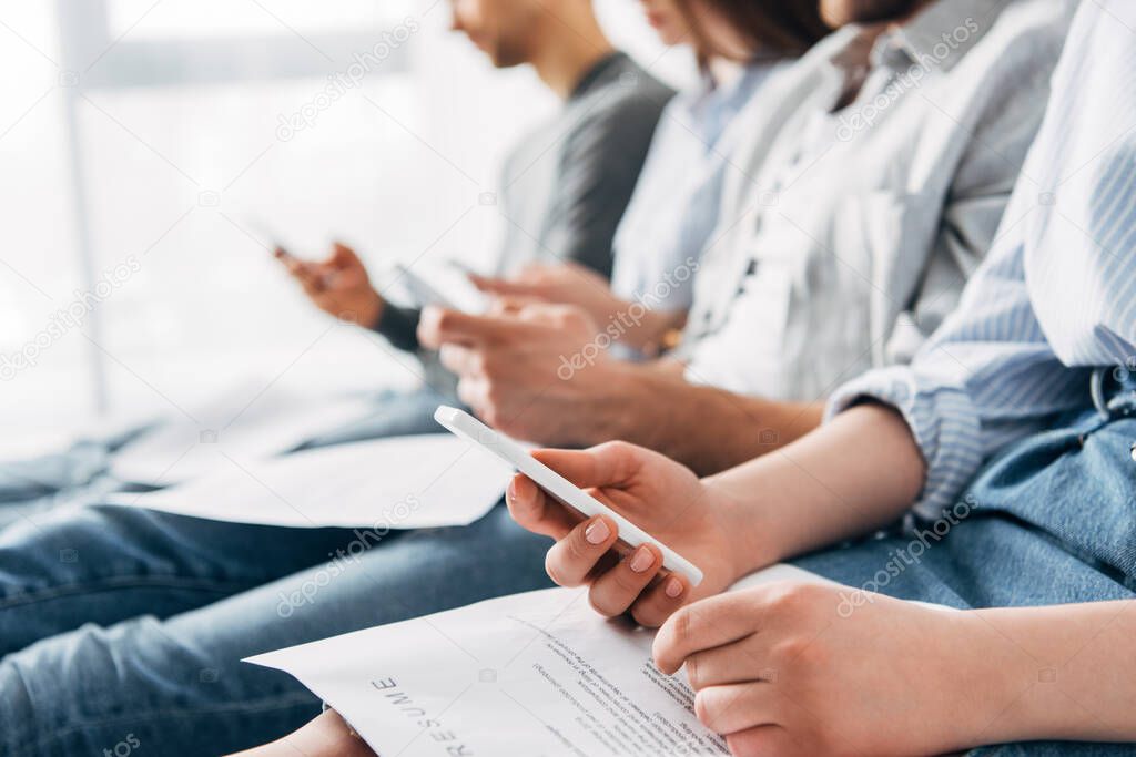 Selective focus of woman with resume using smartphone near applicants in office 