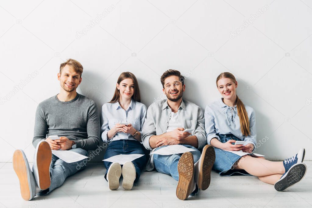 Group of young applicants with resume and smartphones smiling at camera on floor 