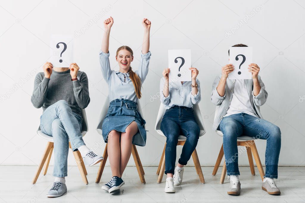 Smiling woman showing yes gesture near employees holding cards with question marks in office 