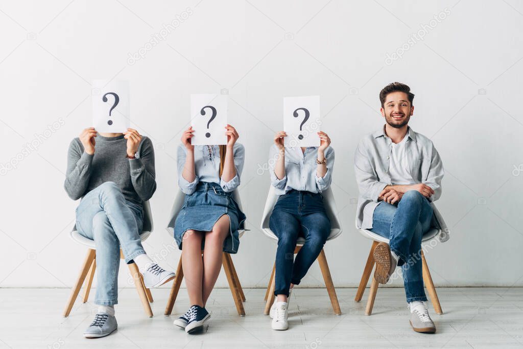Handsome man smiling at camera near employees holding cards with question marks while waiting job interview 