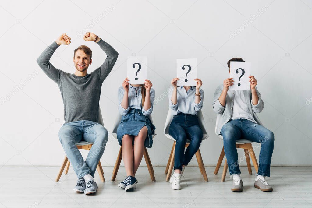 Cheerful man showing yeah gesture near candidates with question marks on cards in office 