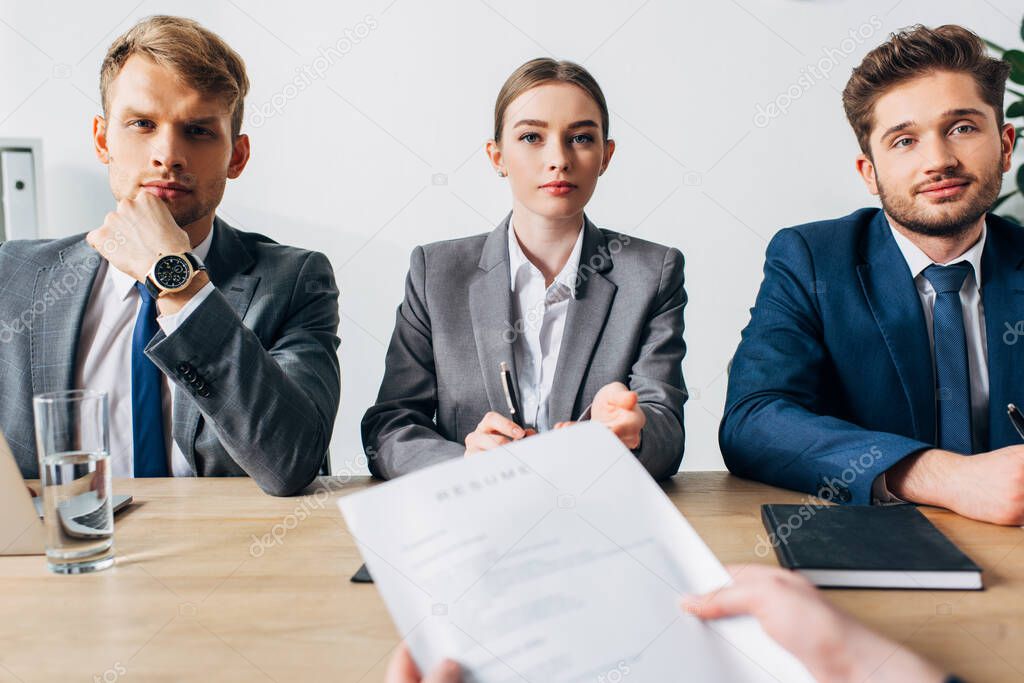 Selective focus of recruiters looking at camera near employee holding resume at table 