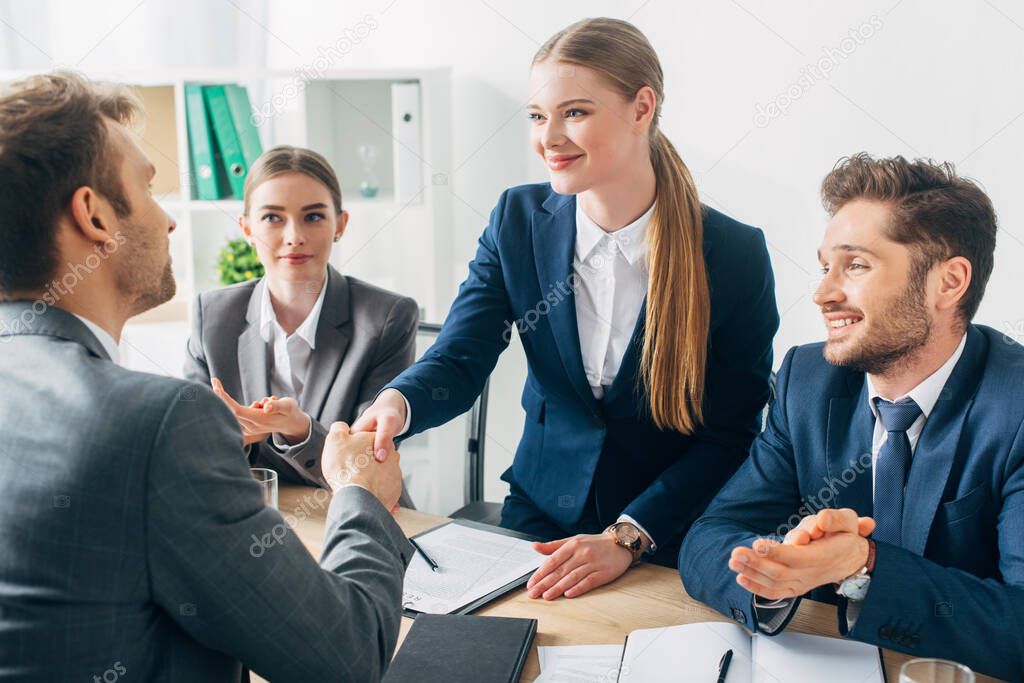 Selective focus of smiling recruiter shaking hands with employee near colleagues clapping at table 