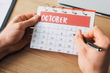 Cropped view of man holding pen and calendar with october month near laptop on table clipart
