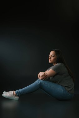 Side view of beautiful sad plus size girl sitting sadly on black background clipart