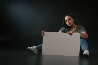 Plus size girl sitting with blank placard on black background clipart