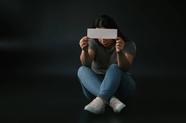 Çapraz bacaklarla oturan ve yüzü siyah arkaplanda boş bir kartla kaplayan büyük boy ön görünüm