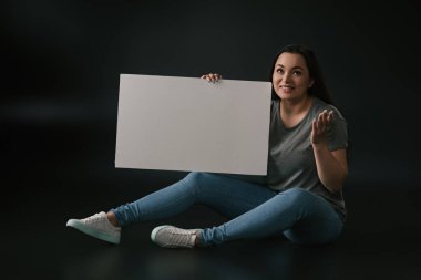 Grateful plus size girl gesturing and sitting with empty board on black background clipart
