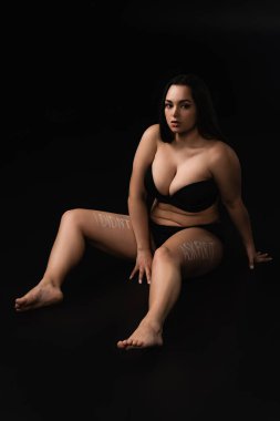 Plus size model in underwear with lettering I Did not Ask For It on body sitting isolated on black  clipart