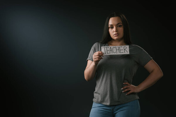 Вид спереди красивой девочки plus size, держащей карточку с учителем слова на чёрном фоне
