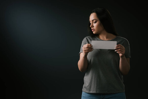 Front view of sad plus size girl holding empty card on black background