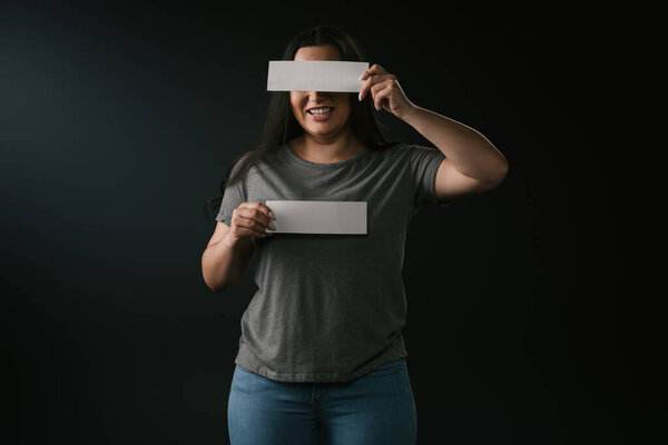 Front view of plus size smiling girl covering eyes and holding empty cards on black background 