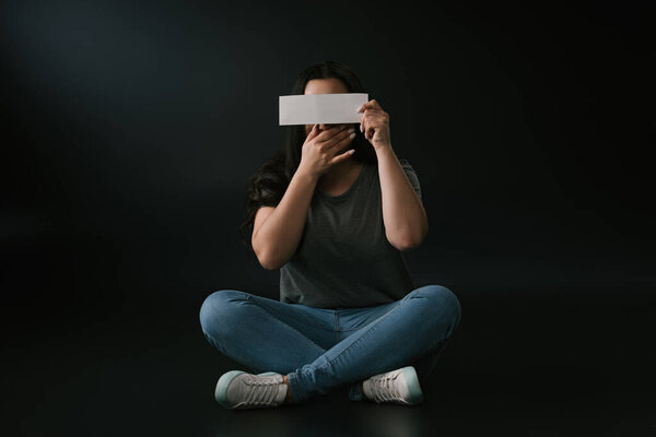 Вид спереди девушки plus size, закрывающей лицо рукой и пустой карточкой и сидящей на чёрном фоне
