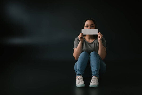 Вид спереди девочки plus size, закрывающей рот пустой карточкой и сидящей на чёрном фоне
