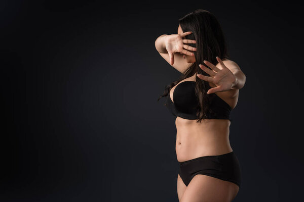 Plus size girl showing stop gesture and covering face with hand on black background 