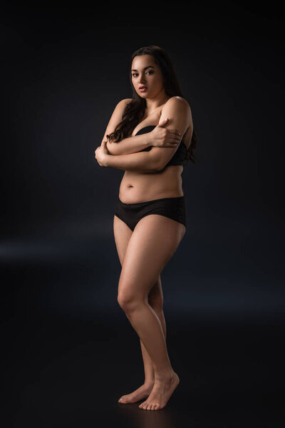 Full length view of plus size girl hugging herself on black background