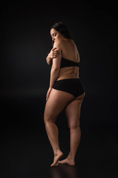 Full length view of plus size girl hiding body with shame on black background