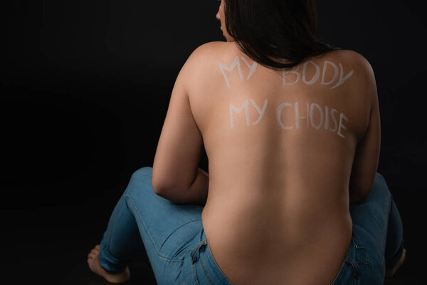 Back view of plus size model with lettering My Body My Choice on naked back sitting in jeans on black background