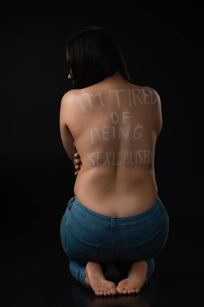 Back view of plus size model hugging her naked back with lettering I Am Tired of Being Sexualised and sitting on black background