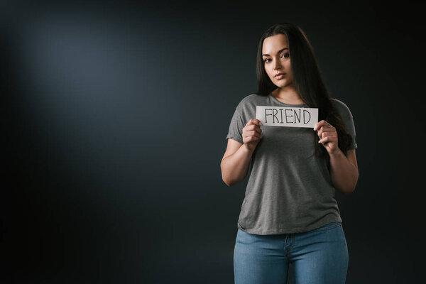 Front view of brunette plus size girl holding card with word friend on black background