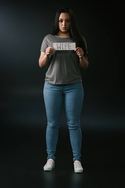 Вид спереди серьёзной девушки plus size, держащей карточку со словом жена на чёрном фоне
