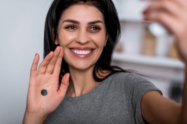 selective focus of smiling woman with bruise on face and black dot on palm taking selfie at home, domestic violence concept  clipart