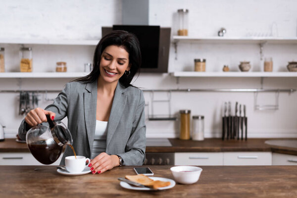 smiling businesswoman pouring coffee in cup near smartphone with blank screen while hiding problem of domestic violence 