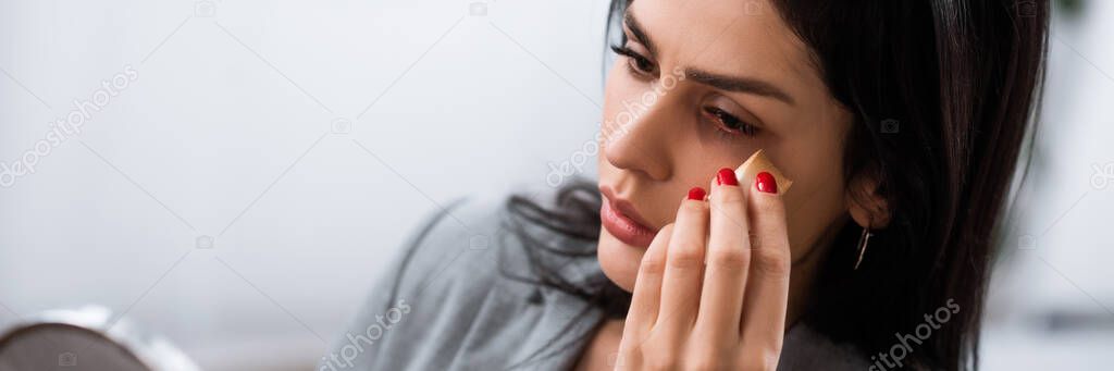 panoramic orientation of businesswoman with bruise on face applying makeup foundation near mirror, domestic violence concept 