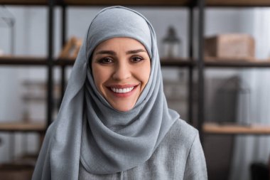 smiling muslim woman in hijab looking at camera in living room, domestic violence concept   clipart