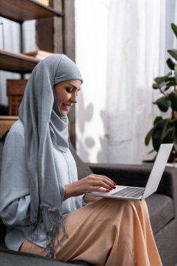 Gülümseyen Arap kadın oturma odasında dizüstü bilgisayar kullanıyor. 