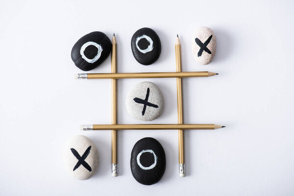 top view of tic tac toe game with grid made of pencils, and pebbles marked with crosses and naughts on white surface