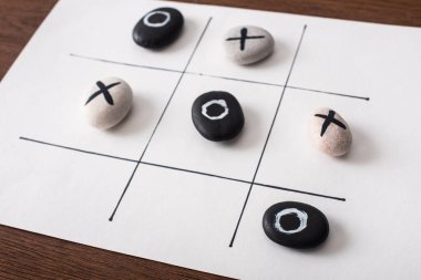 tic tac toe game on white paper with pebbles marked with naughts and crosses on wooden surface clipart