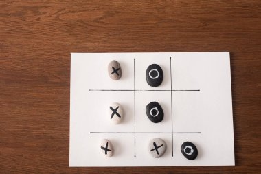 top view of tic tac toe game on white paper with stones marked with naughts and crosses on wooden surface clipart
