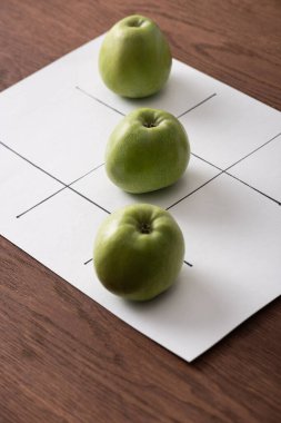 Tahta yüzey üzerinde üç yeşil elma sırasıyla beyaz kağıt üzerinde tic tac toe oyunu