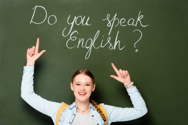 smiling girl with backpack pointing at chalkboard with do you speak English lettering clipart