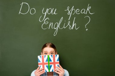 pretty girl with obscure face holding book with uk flag near chalkboard with do you speak English lettering clipart