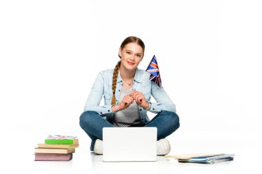smiling girl sitting on floor with laptop, books and copybooks and holding uk flag isolated on white clipart