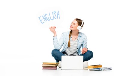 smiling girl sitting on floor in headphones with speech bubble with english lettering near laptop, books and copybooks isolated on white clipart