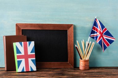 empty chalkboard near pencils, books and uk flag on wooden table near blue wall clipart