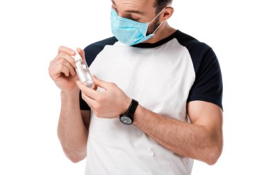 Tıbbi maskeli bir adam elinde sprey şişesiyle beyaza izole edilmiş antibakteriyel sıvı tutuyor. 