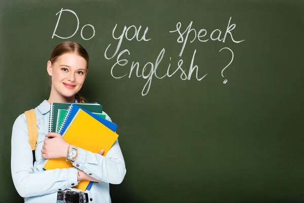 smiling girl with copybooks and backpack standing near chalkboard with do you speak English lettering