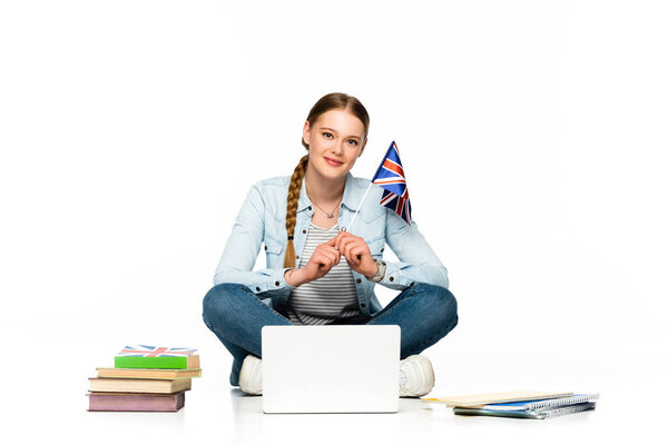 smiling girl sitting on floor with laptop, books and copybooks and holding uk flag isolated on white