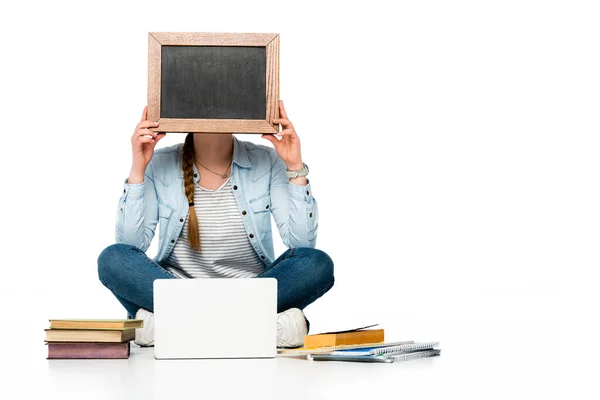 girl sitting on floor with obscure face near laptop, books and copybooks and holding chalkboard isolated on white