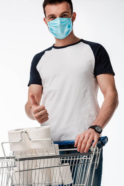 мужчина в медицинской маске показывает большой палец вверх возле корзины с туалетной бумагой, изолированной на белом
