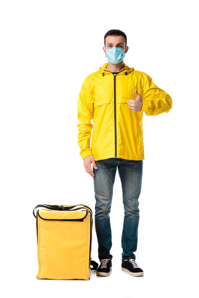 delivery man in medical mask standing near bag with order and showing thumb up isolated on white 