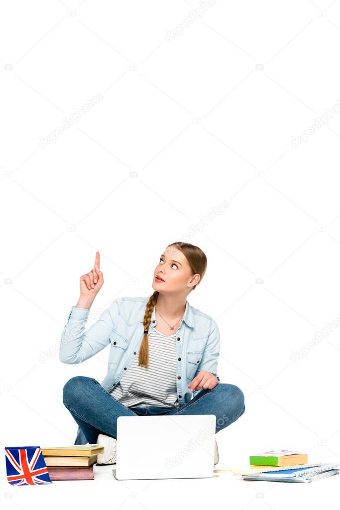 girl sitting on floor with laptop, books and copybooks, uk flag and pointing up isolated on white