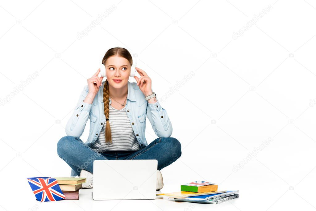 smiling girl sitting on floor in headphones near laptop, books and copybooks, uk flag isolated on white